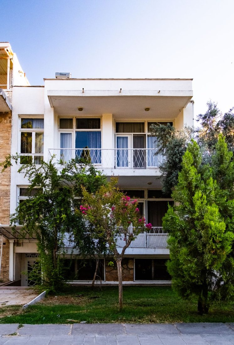 The front of a pale house with balconies and trees.