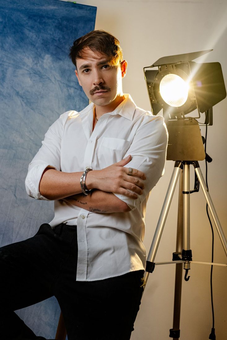 A man seated near a bright studio lamp and backdrop.